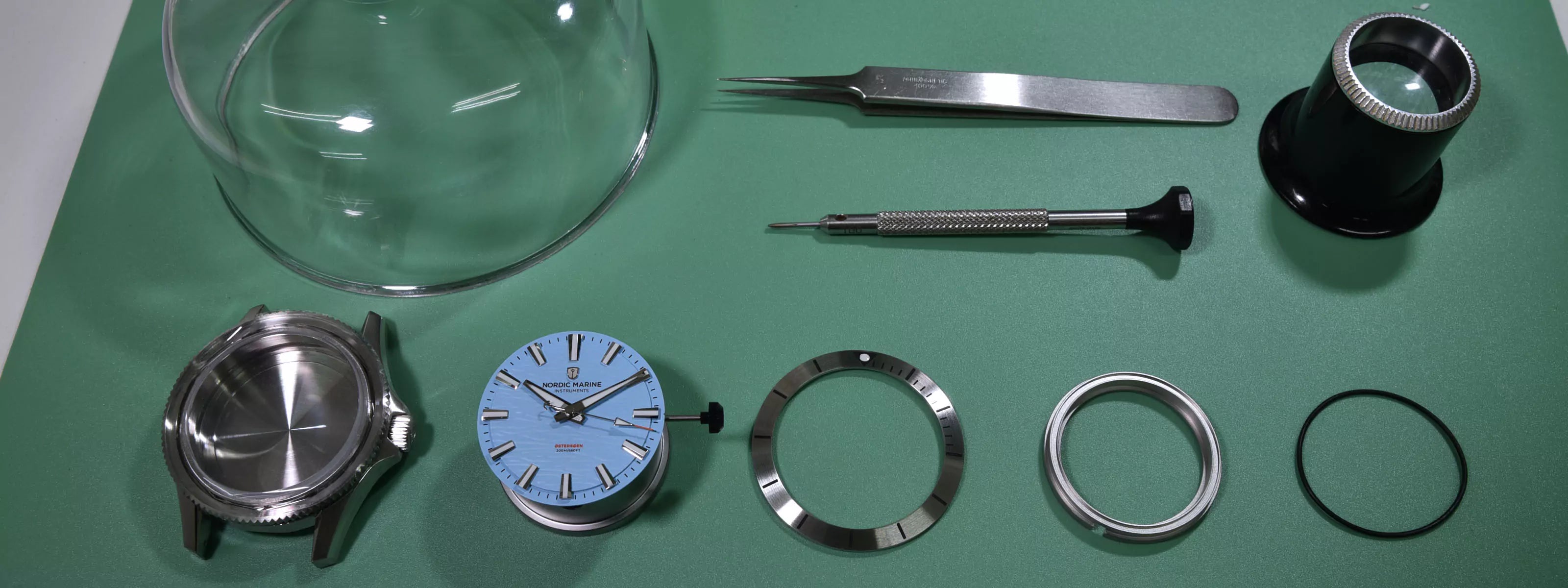 Watchmaker watch parts including steel case, dial glass, bezel, and metal components on a green surface