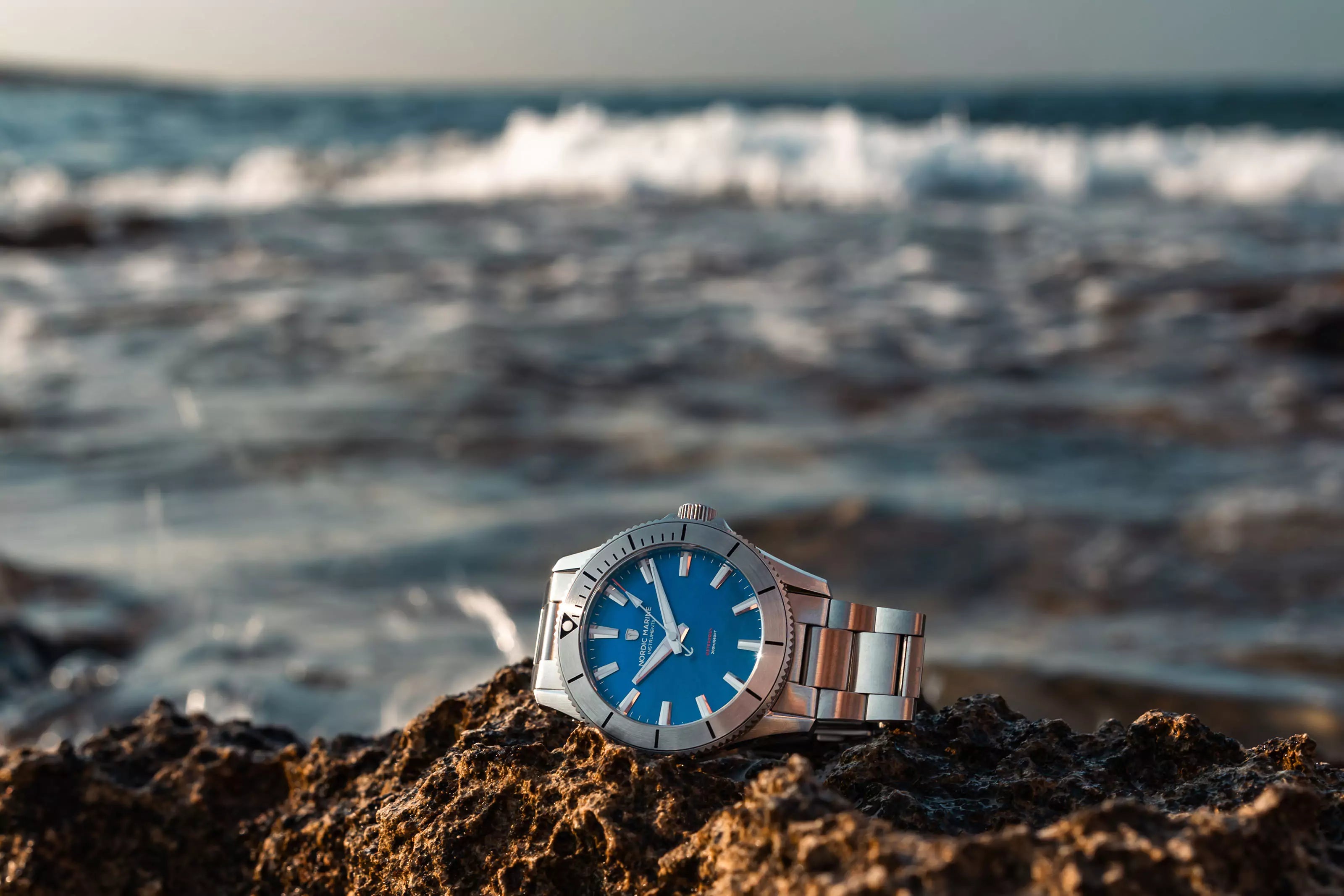 Silver watch with blue mother-of-pearl dial on a rock by the ocean with waves